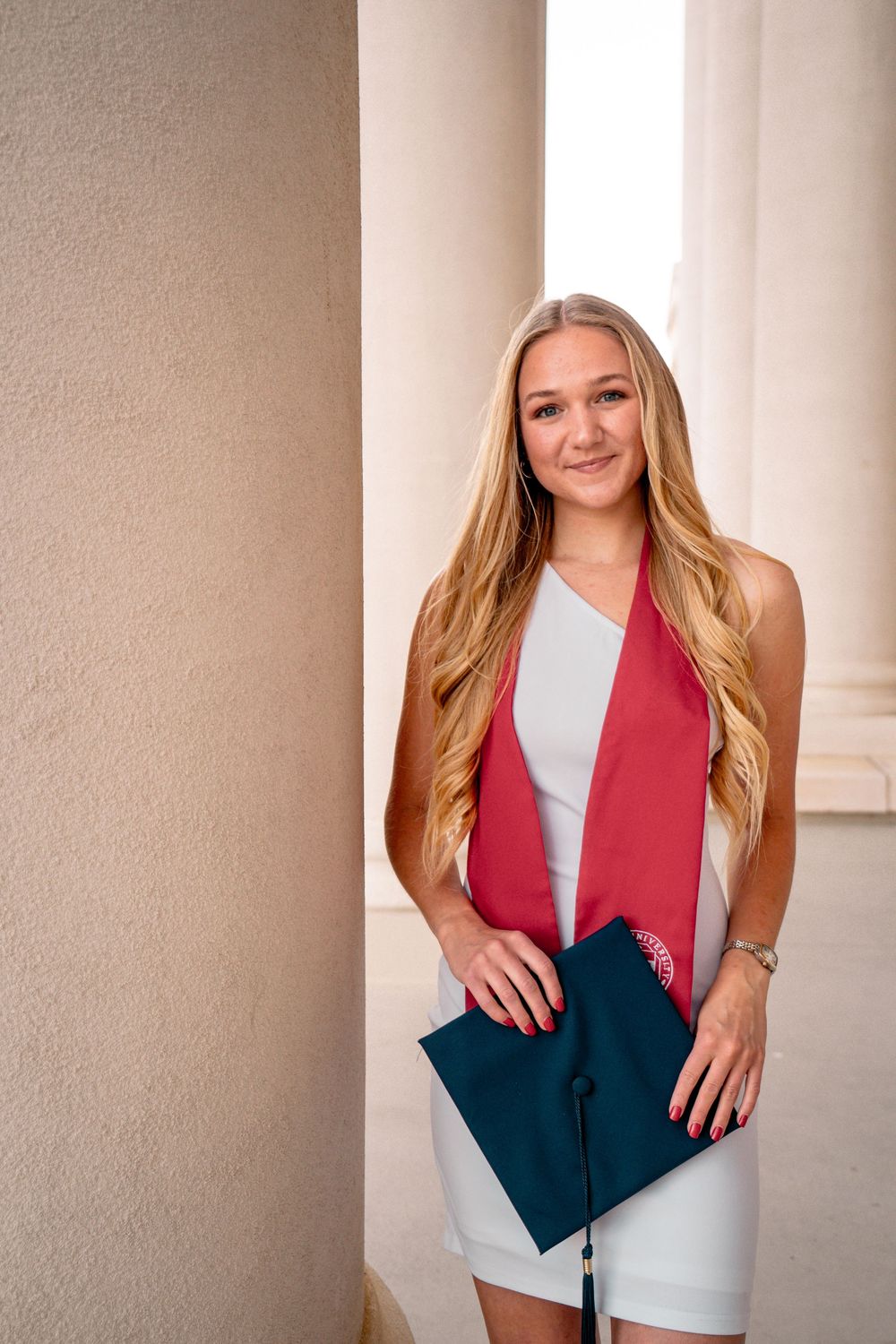 Young woman in white dress with red graduation sash holds cap between columns for senior portrait.