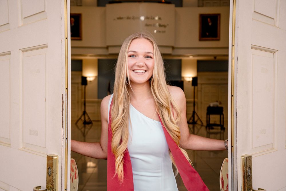 Smiling young woman with long blonde hair in white dress standing in elegant doorway with gold handles.