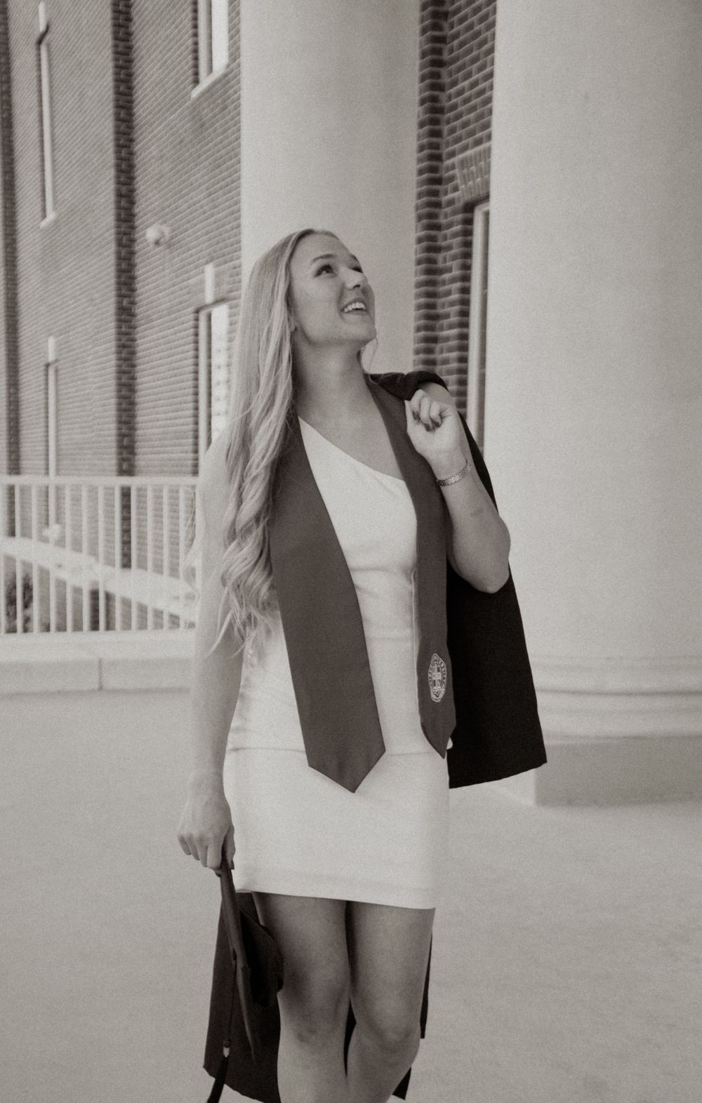 Young woman in white dress with graduation stole and cap poses outdoors near brick building in black and white photo.