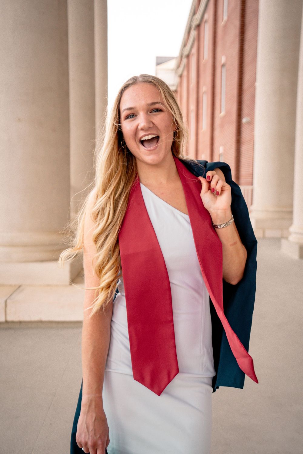 Smiling blonde woman in white dress with red graduation stole and navy gown over shoulder, posing by columns.