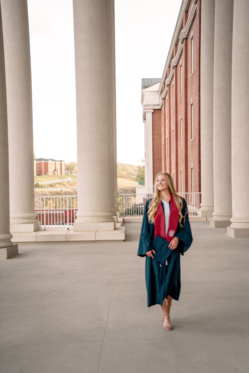Young woman in teal graduation gown and red sash walks between columns of a brick university building.