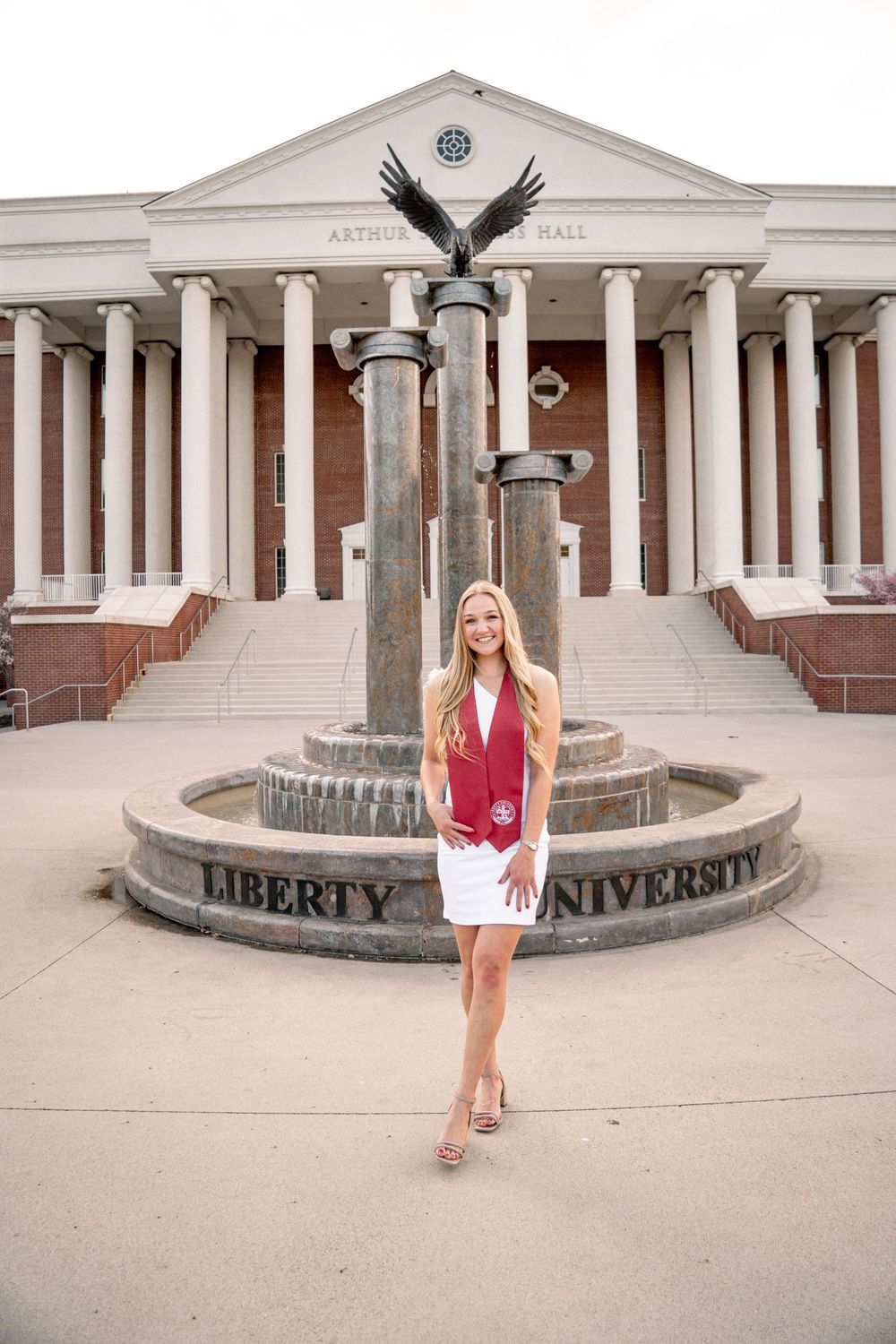 Liberty University graduate smiles in white dress and red stole, posing by the eagle fountain outside DeMoss Hall.