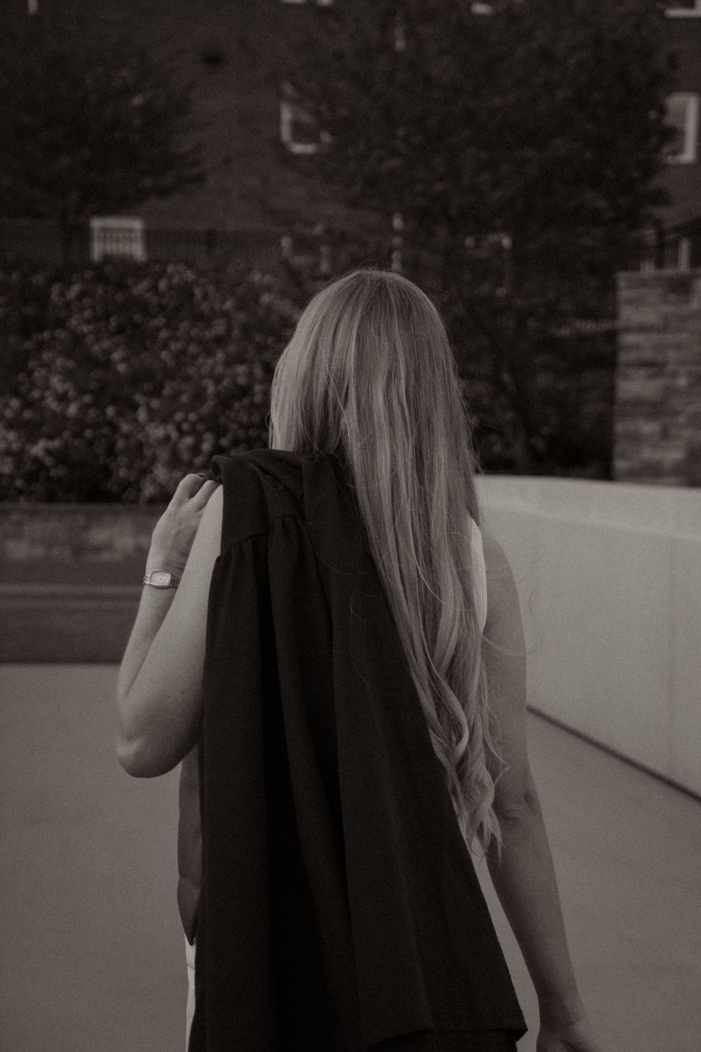 Black and white photo of a blonde woman from behind, wearing a black top, standing outdoors near trees.