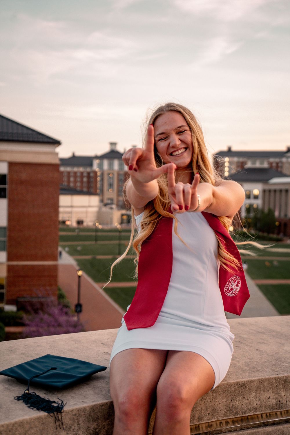 Smiling female graduate in white dress with red sash makes hand gestures on campus at sunset.