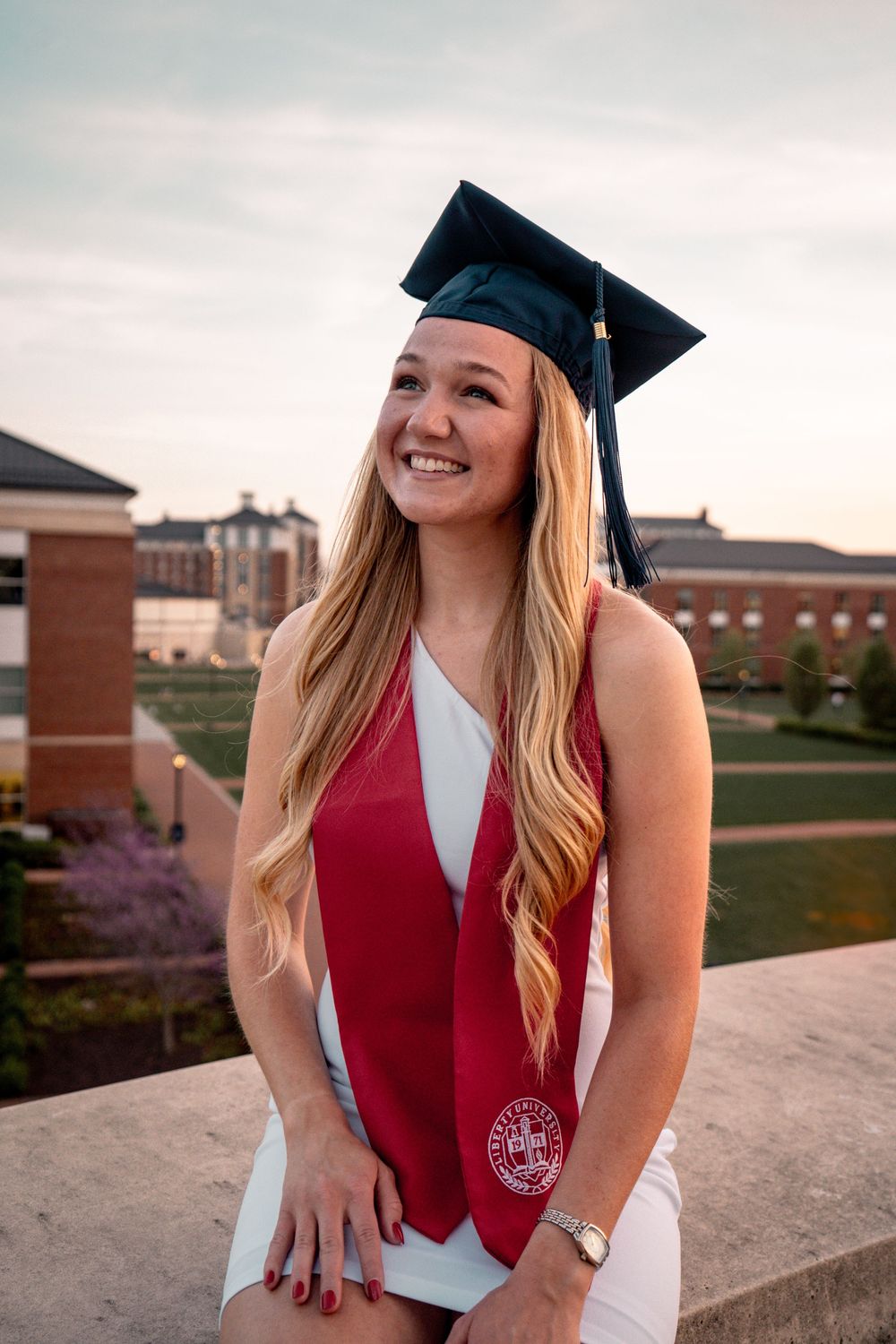 Smiling young woman in graduation cap and red stole posing outdoors on a college campus at sunset.