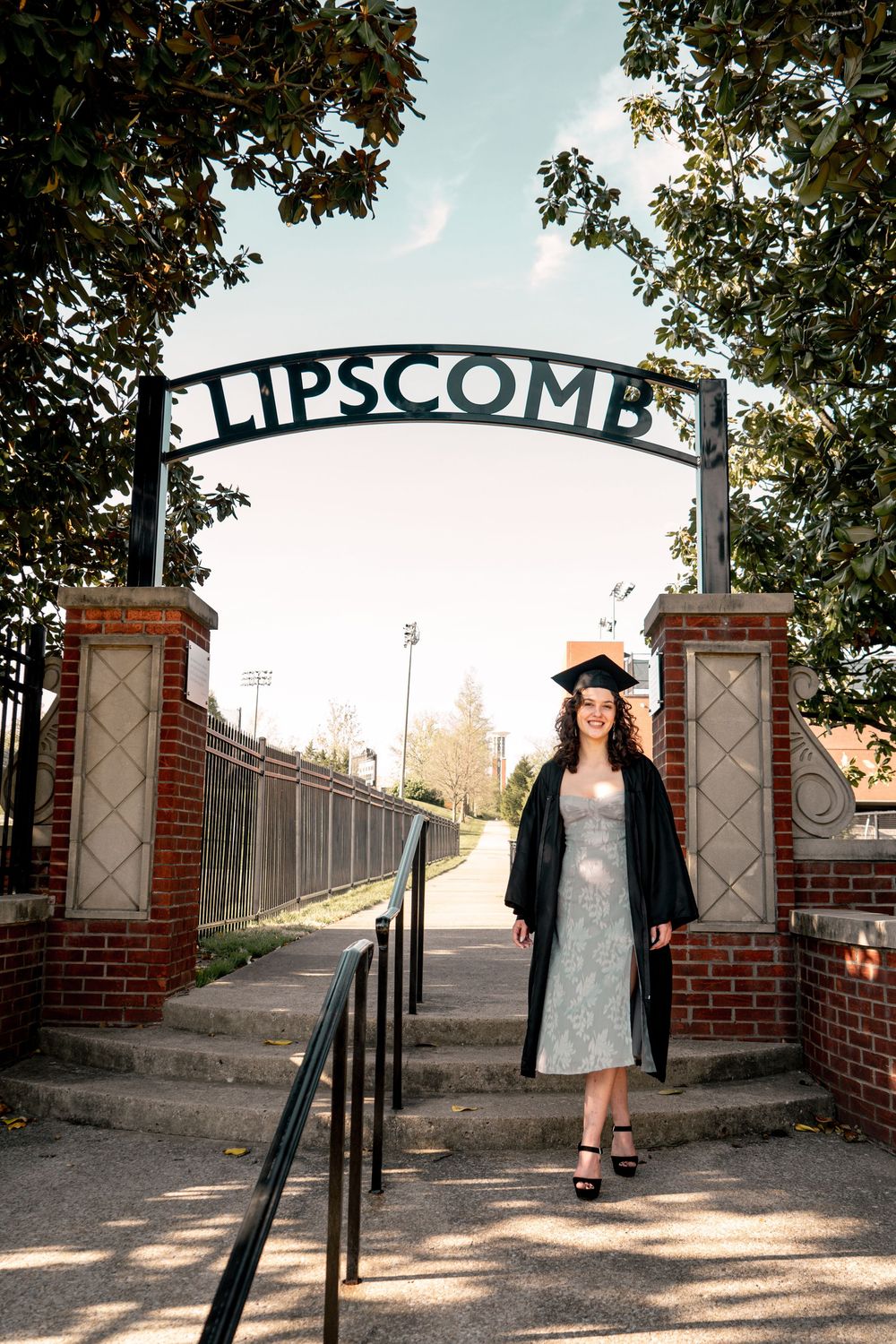 Graduate in cap and gown poses under Lipscomb University arch on a sunny day.