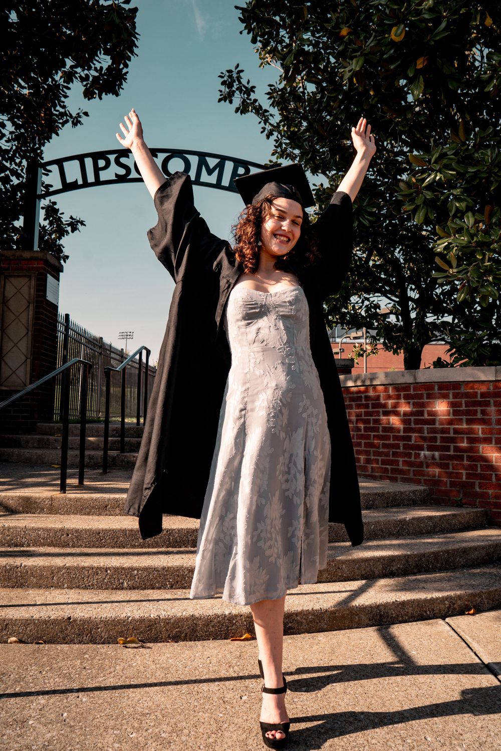 A joyful graduate in cap and gown raises arms in celebration on steps outside Lipscomb University.