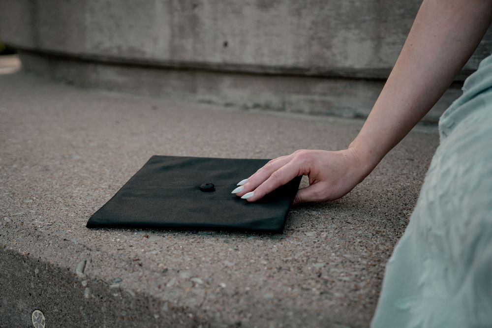 A graduate's hand resting on a black mortarboard cap placed on concrete steps, wearing a light blue dress.