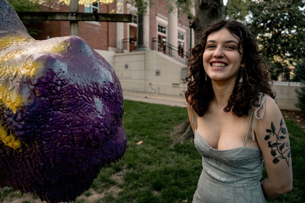 Smiling woman with curly hair and floral tattoo poses beside colorful abstract sculpture outdoors on college campus.