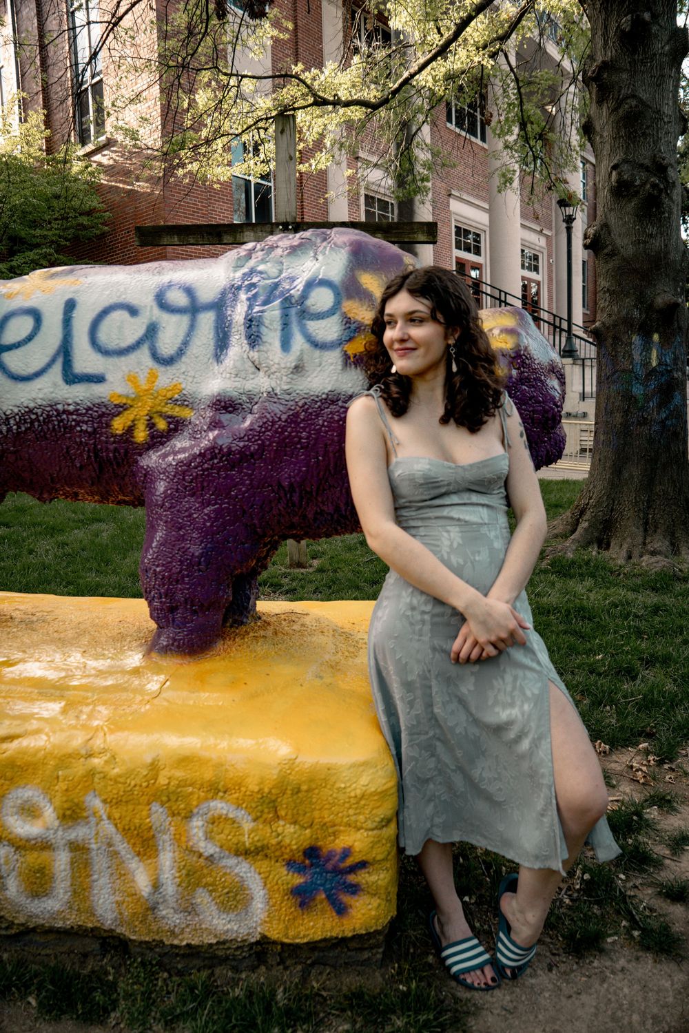 Young woman in silver dress poses beside colorful graffiti bear sculpture on yellow base outdoors.