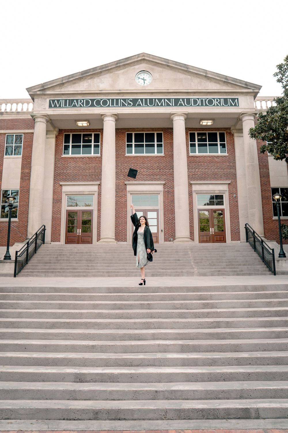 Graduate tosses cap on steps of Willard Collins Alumni Auditorium in academic regalia during commencement celebration.