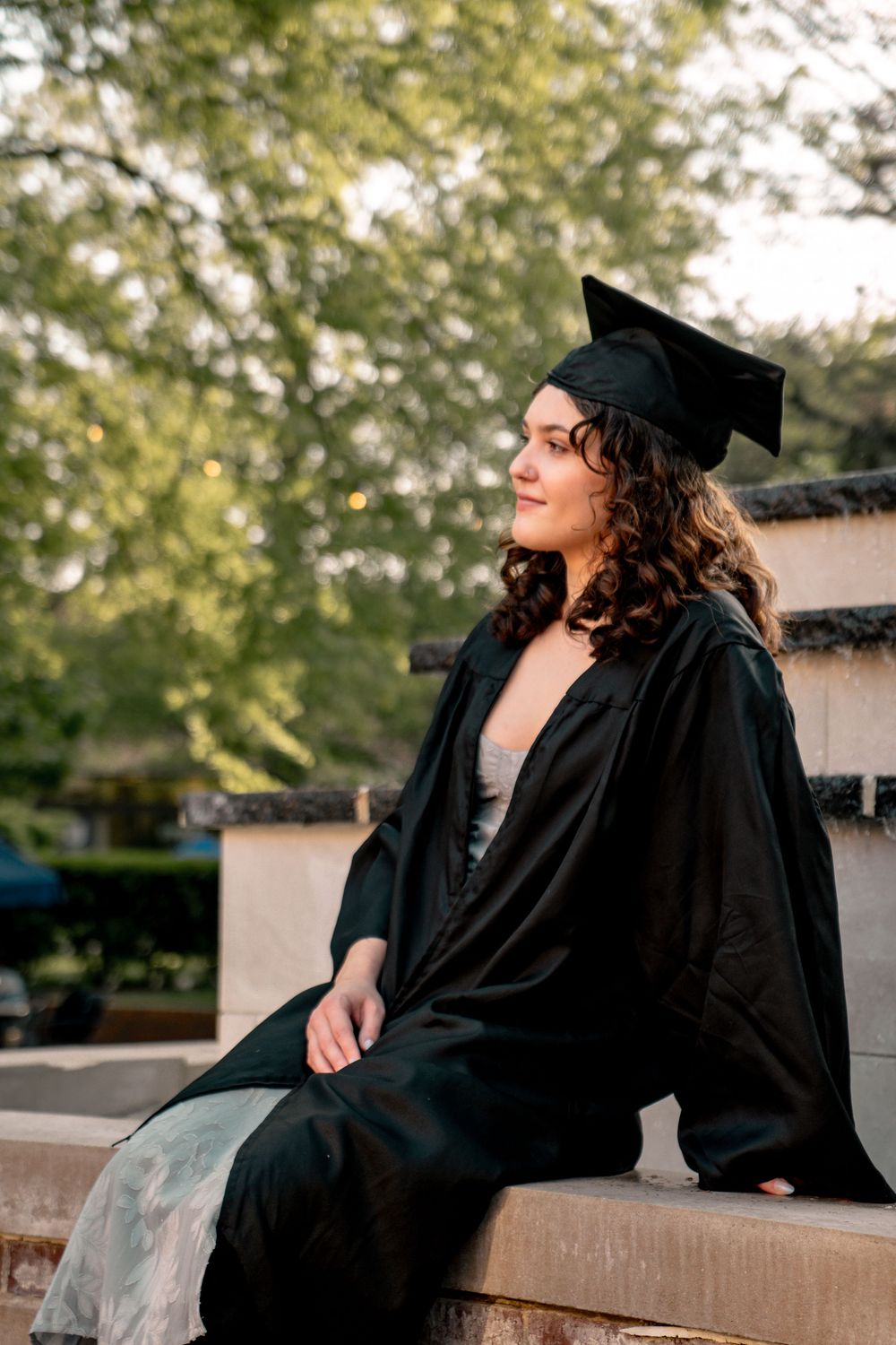 Young woman in black graduation cap and gown sitting outdoors, smiling thoughtfully with green trees in background.