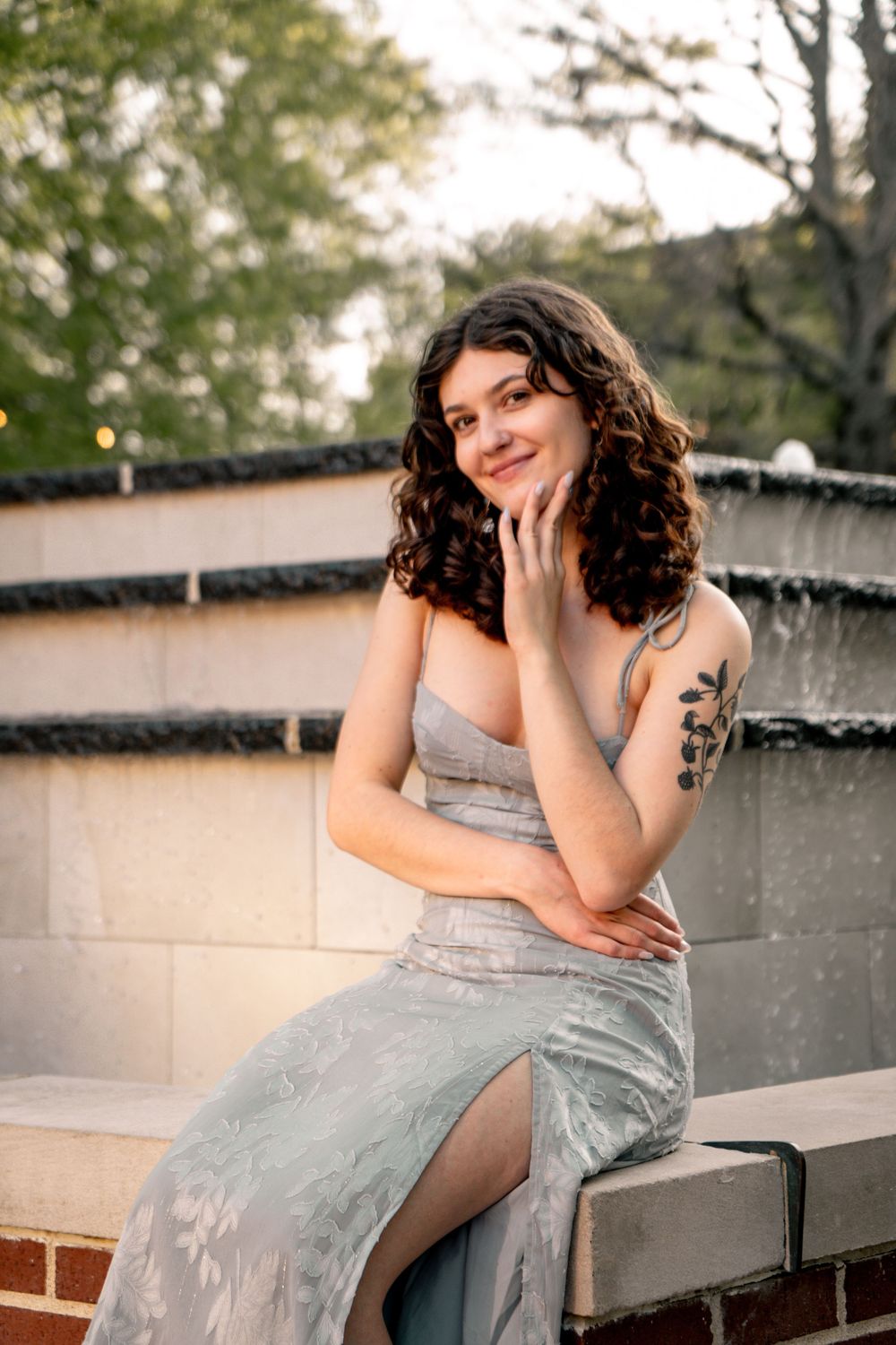 Young woman in silver gown with floral tattoo sits by fountain, smiling elegantly at outdoor evening event.