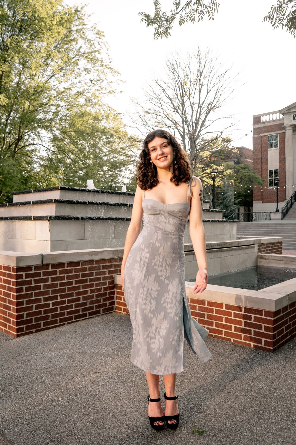 Young woman in silver midi dress and black heels smiling outdoors near brick fountain and trees.