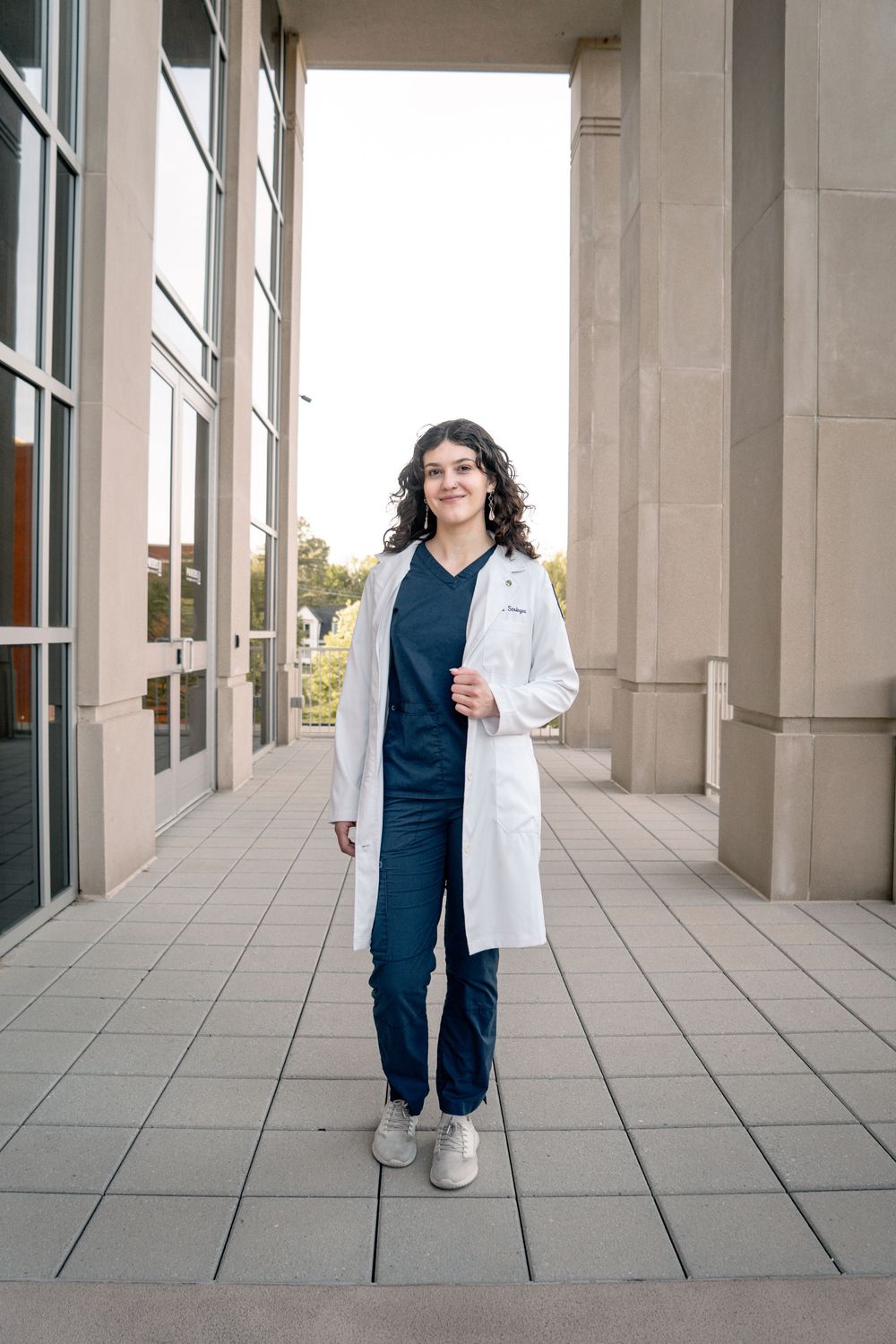 A young female medical professional in white lab coat and navy scrubs stands confidently outside a modern building.