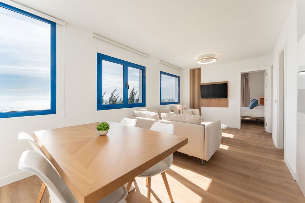 Modern open-plan living and dining room with blue-framed windows overlooking the sea, light wood flooring, and white furniture.