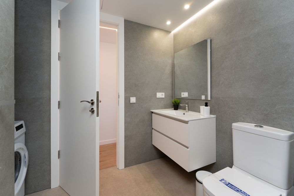 Modern bathroom with gray walls, white floating vanity, mirror with LED lighting, toilet, and visible washing machine.