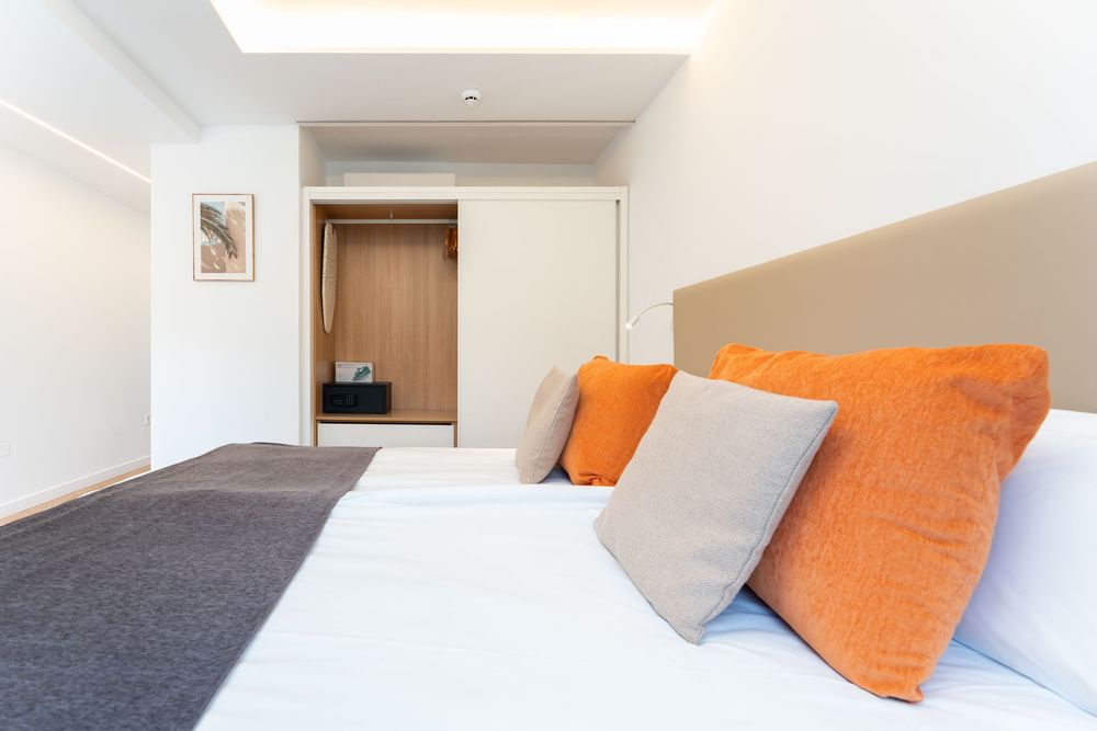 Modern bedroom with orange and gray pillows on a white bed, featuring a wooden wardrobe and recessed lighting.