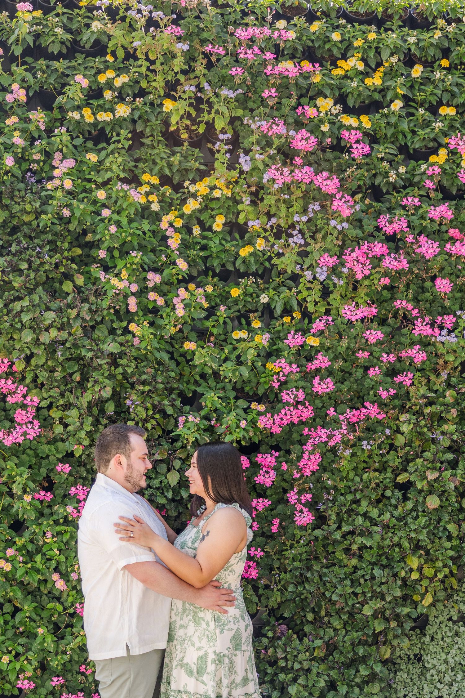Couple embracing and smiling at each other in front of a lush green wall covered in colorful pink and yellow flowers.