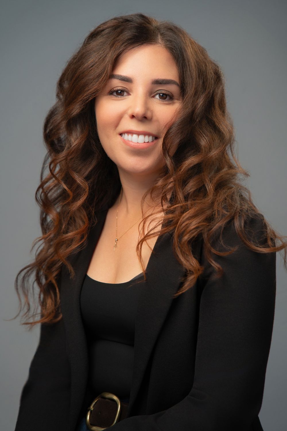 Professional headshot of a smiling woman with curly brown hair wearing a black blazer and gold necklace.