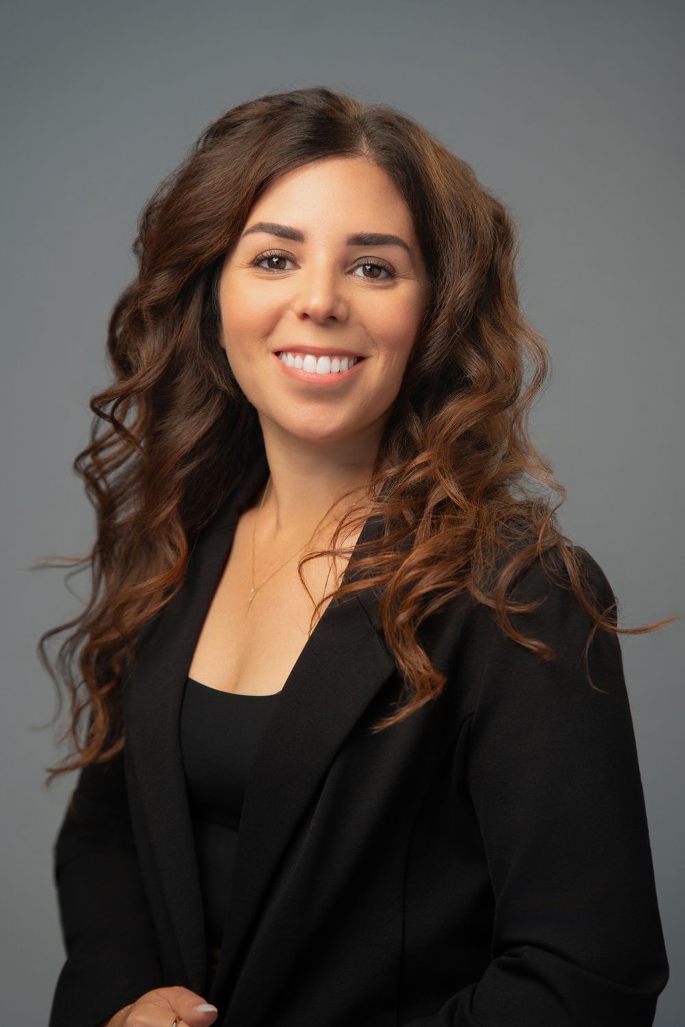 Professional headshot of a smiling woman with curly brown hair wearing a black blazer against a gray background.