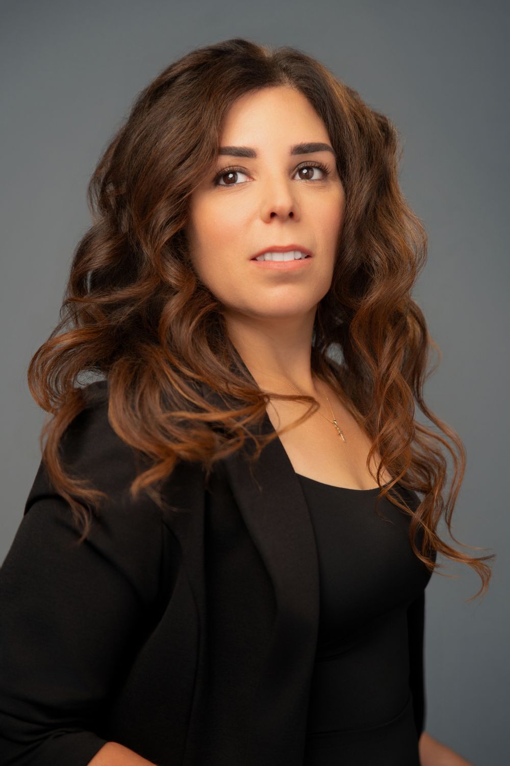 Professional headshot of a woman with curly brown hair wearing a black blazer against a gray background.