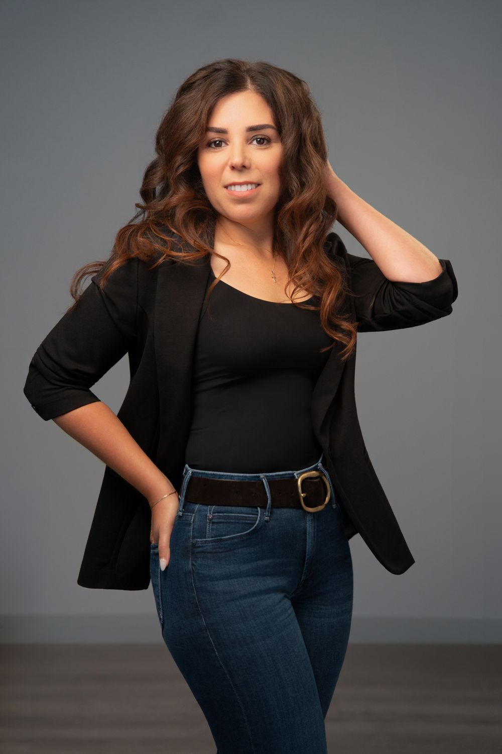 Woman in black blazer and jeans posing confidently with hand in hair against gray studio background.