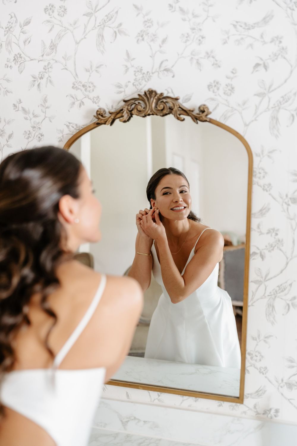 Smiling bride in white dress adjusting earrings while looking in an ornate gold mirror.