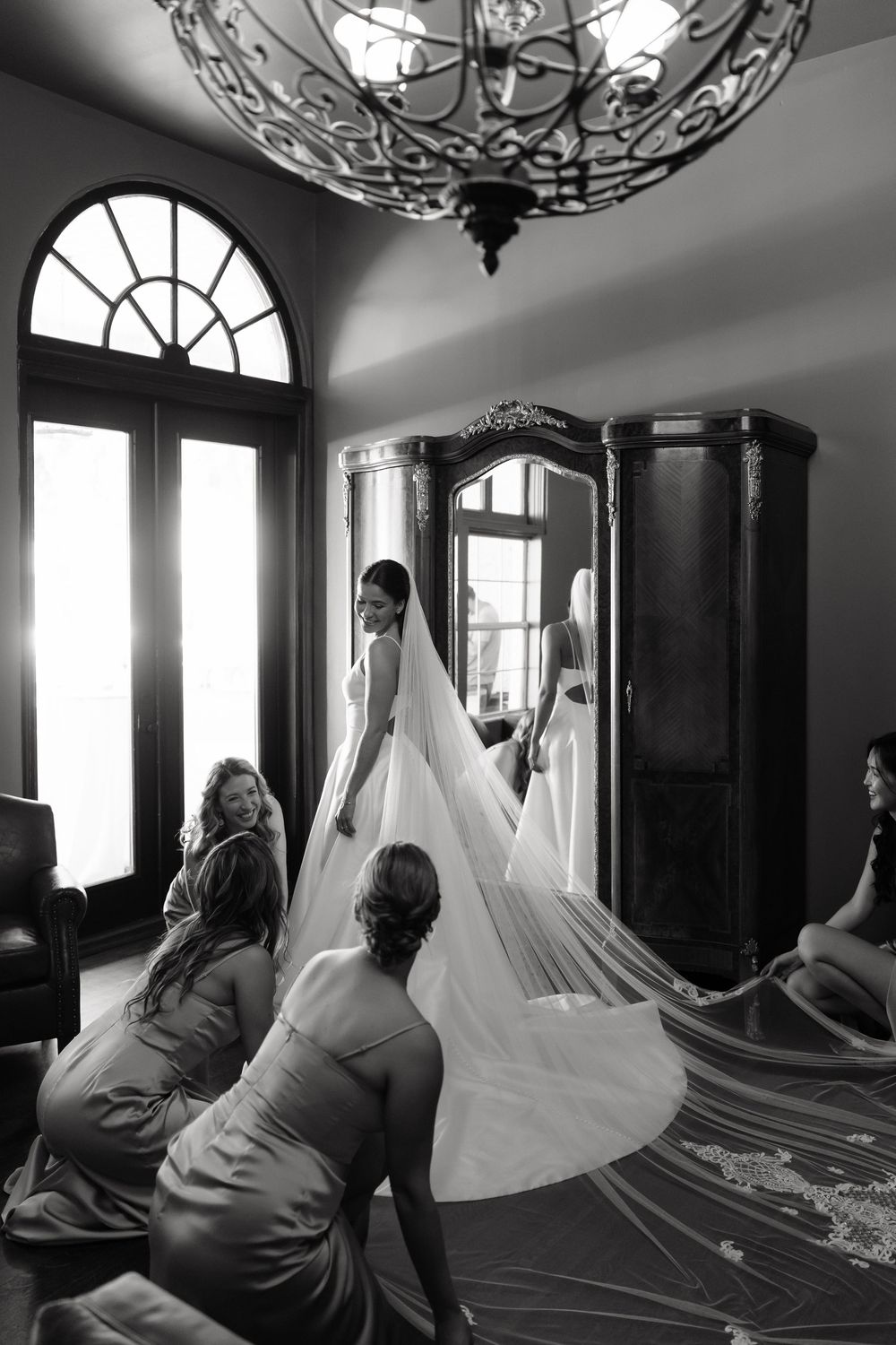 Bridesmaids help a bride arrange her long lace veil in an elegant room with a vintage mirror.