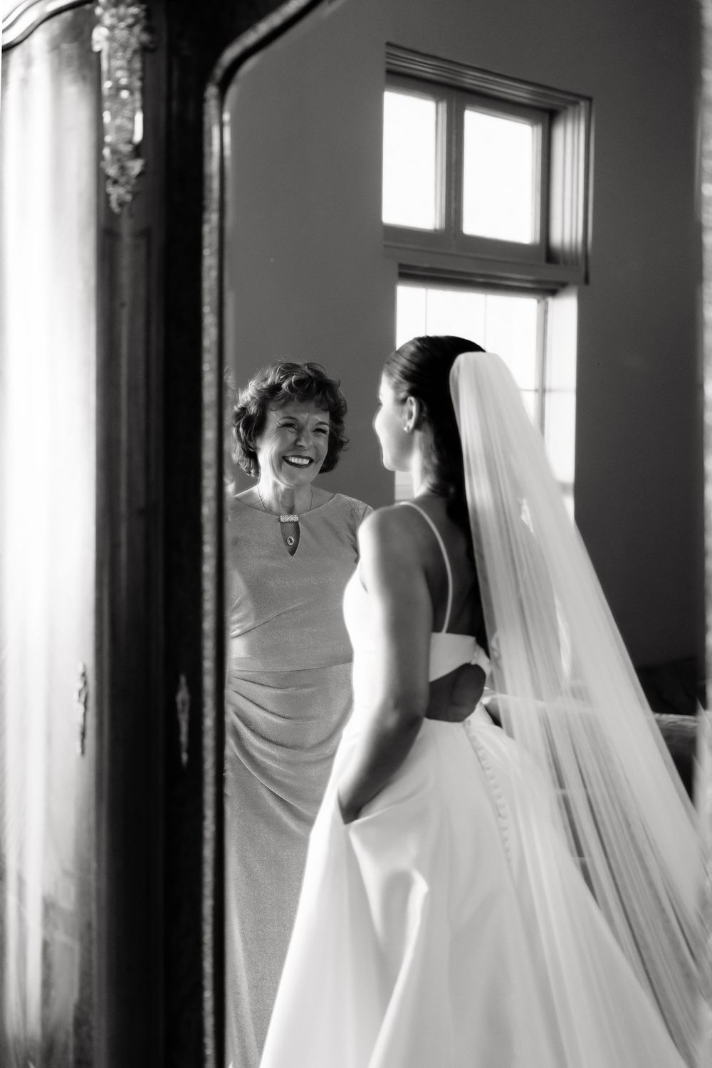 Black and white photo of smiling mother admiring bride in wedding gown before ceremony.