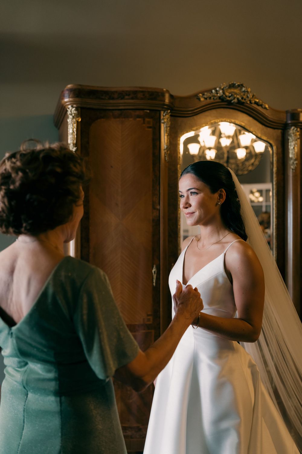 Bride in white gown holds hands with mother in green dress before wedding ceremony near ornate wooden armoire.