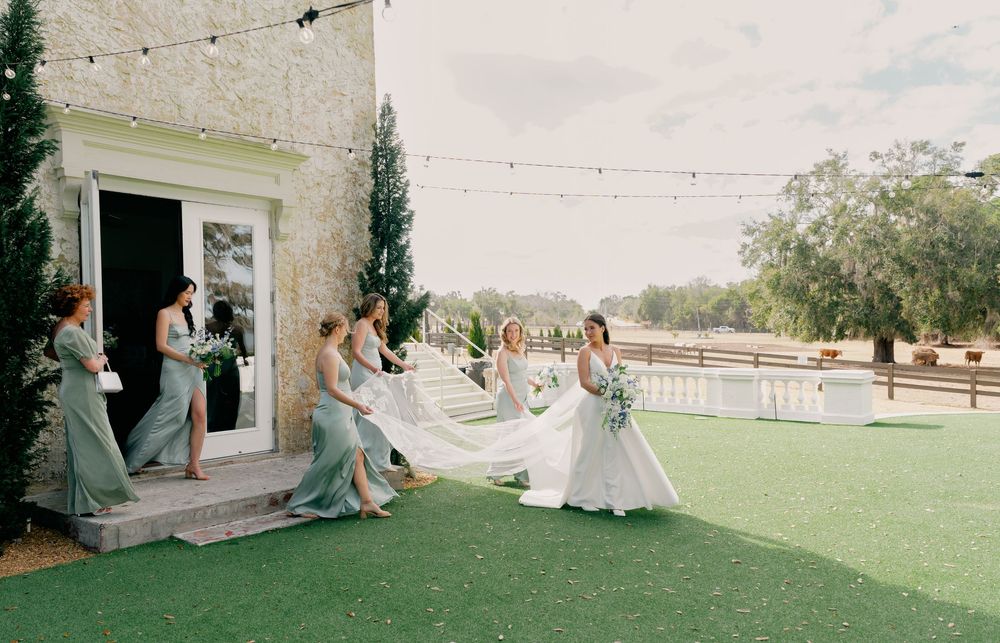 Bride in white gown with bridesmaids in sage green dresses holding veil on green lawn at elegant outdoor wedding venue.