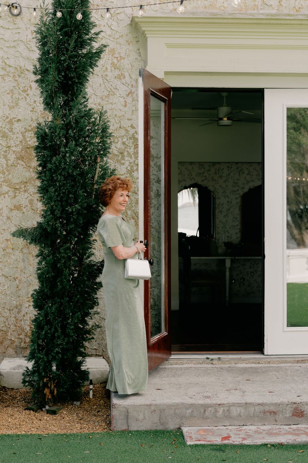 Elegant woman in sage green dress holding white handbag standing at venue entrance beside tall cypress tree.