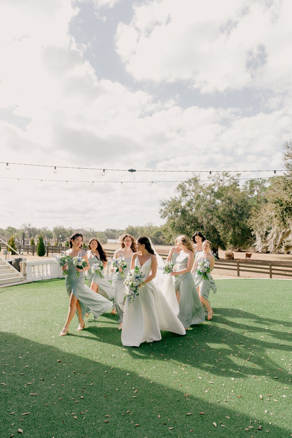 Bride and bridesmaids in sage green gowns walking outdoors on green lawn under string lights.
