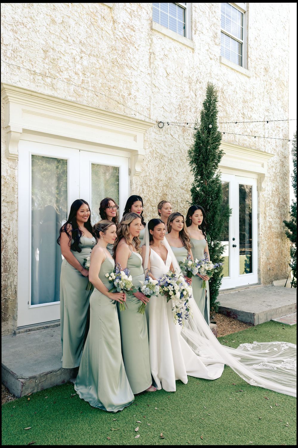 Bride in white gown poses with bridesmaids in sage green dresses outside elegant stone venue.