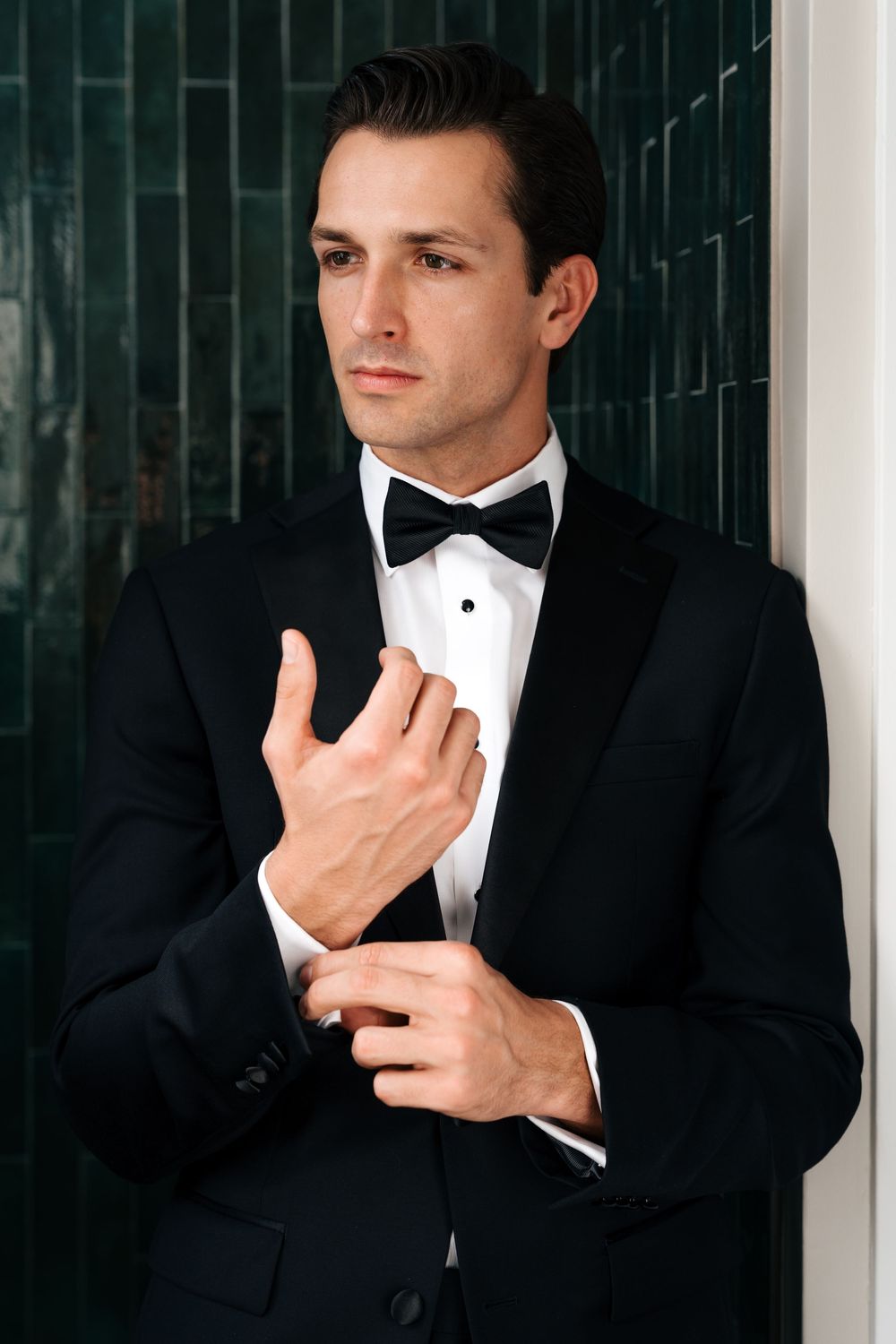 Elegant groom in black tuxedo with bow tie adjusting cufflinks against green tiled wall.