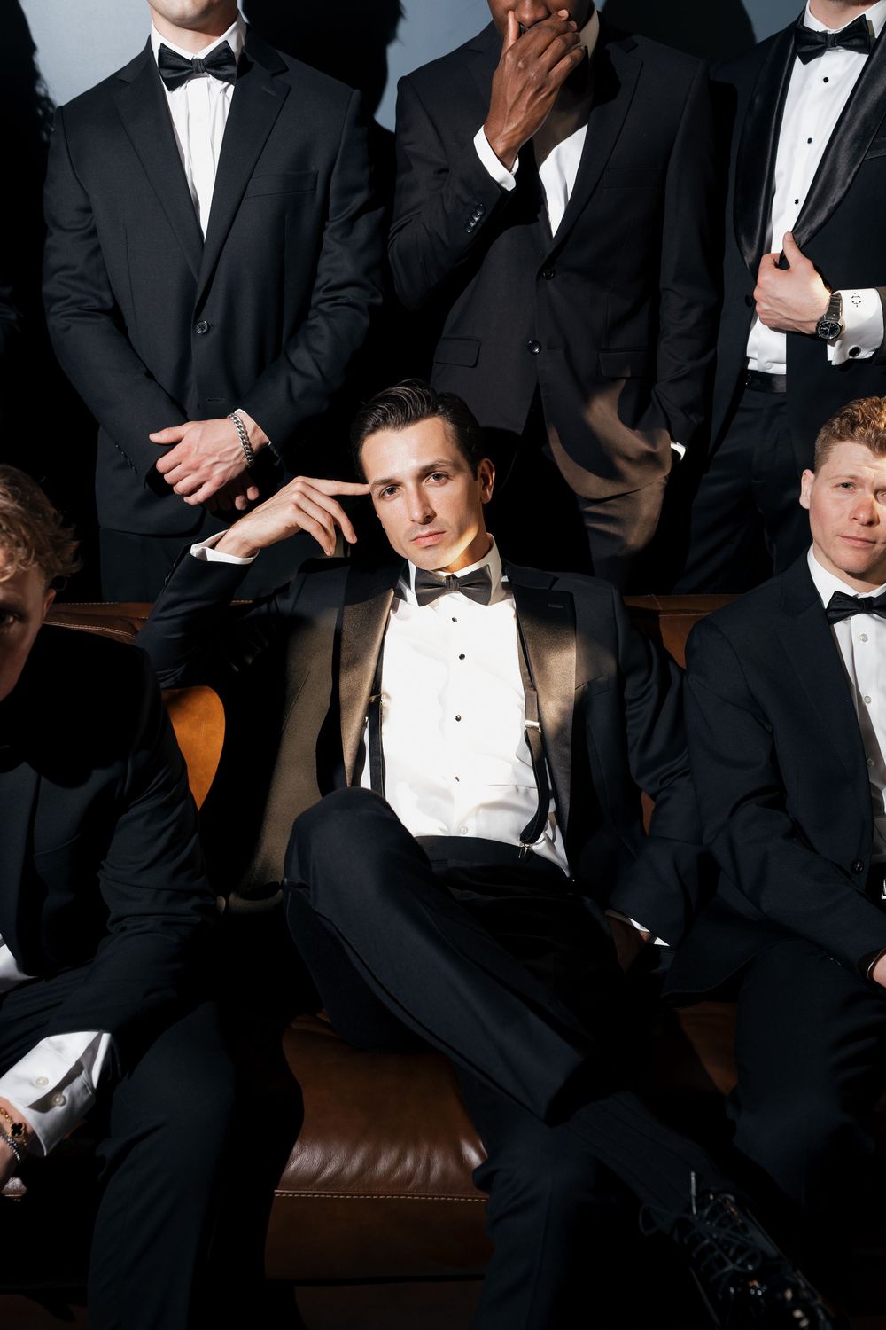 Group of groomsmen in black tuxedos posing together with groom seated on leather sofa.