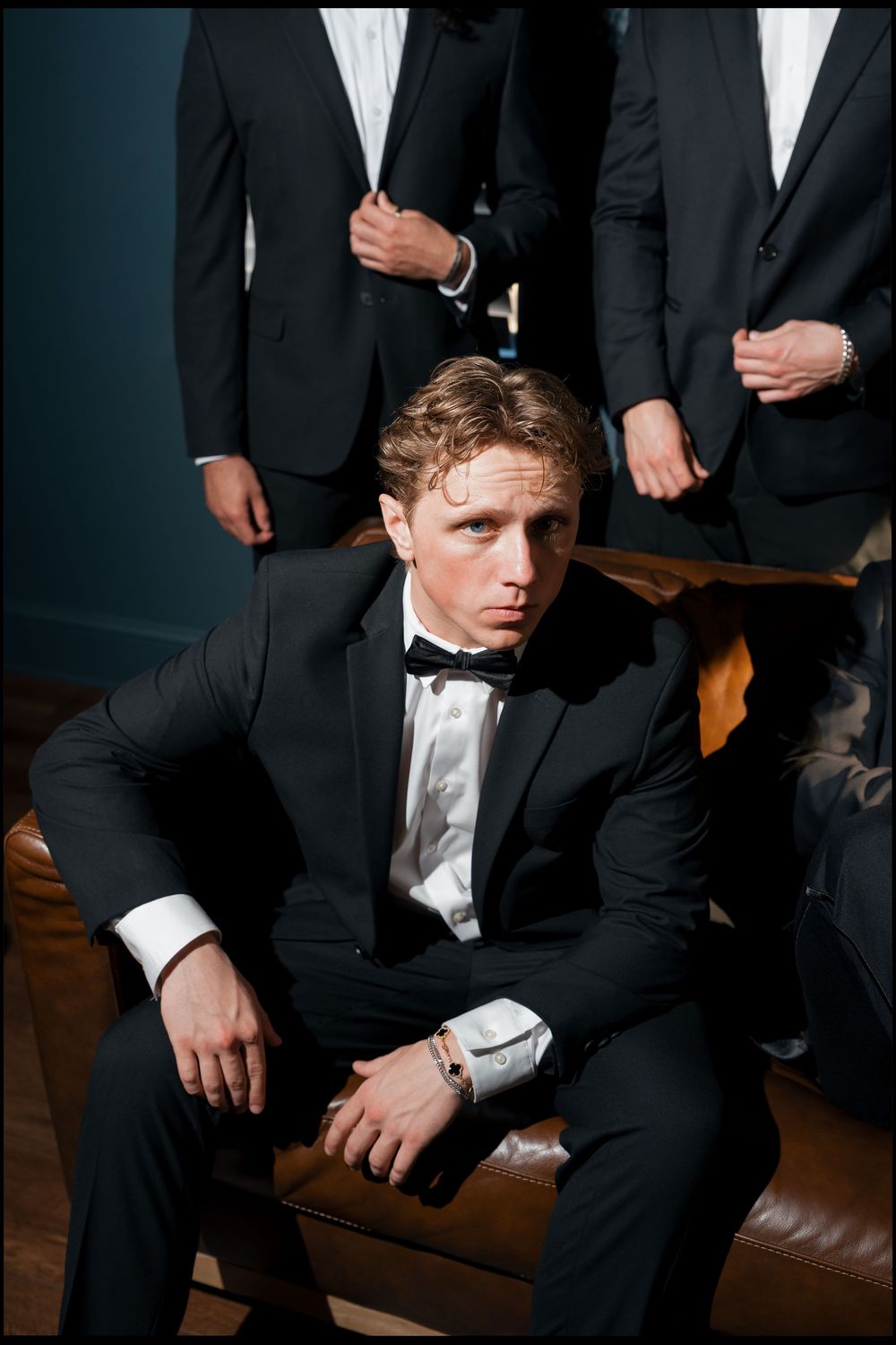 Groom in black tuxedo with bow tie sits confidently with groomsmen standing behind him.