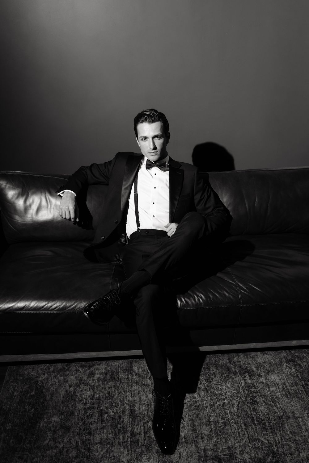 A groom in a black tuxedo and bow tie sits confidently on a leather sofa in dramatic lighting.