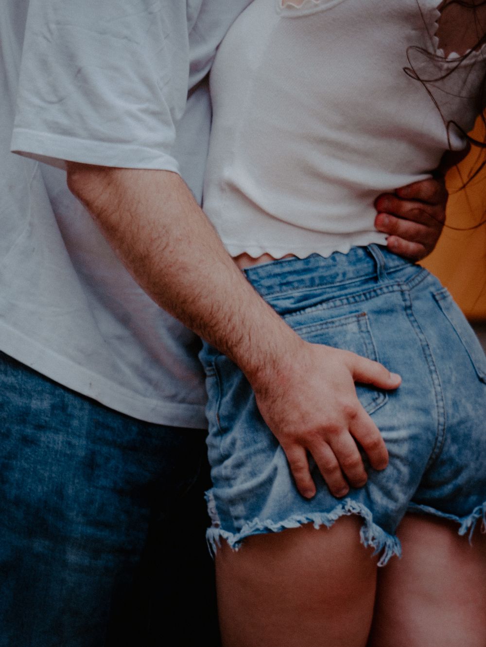 Couple in casual denim and white outfits standing close together, man's hand resting on woman's hip.
