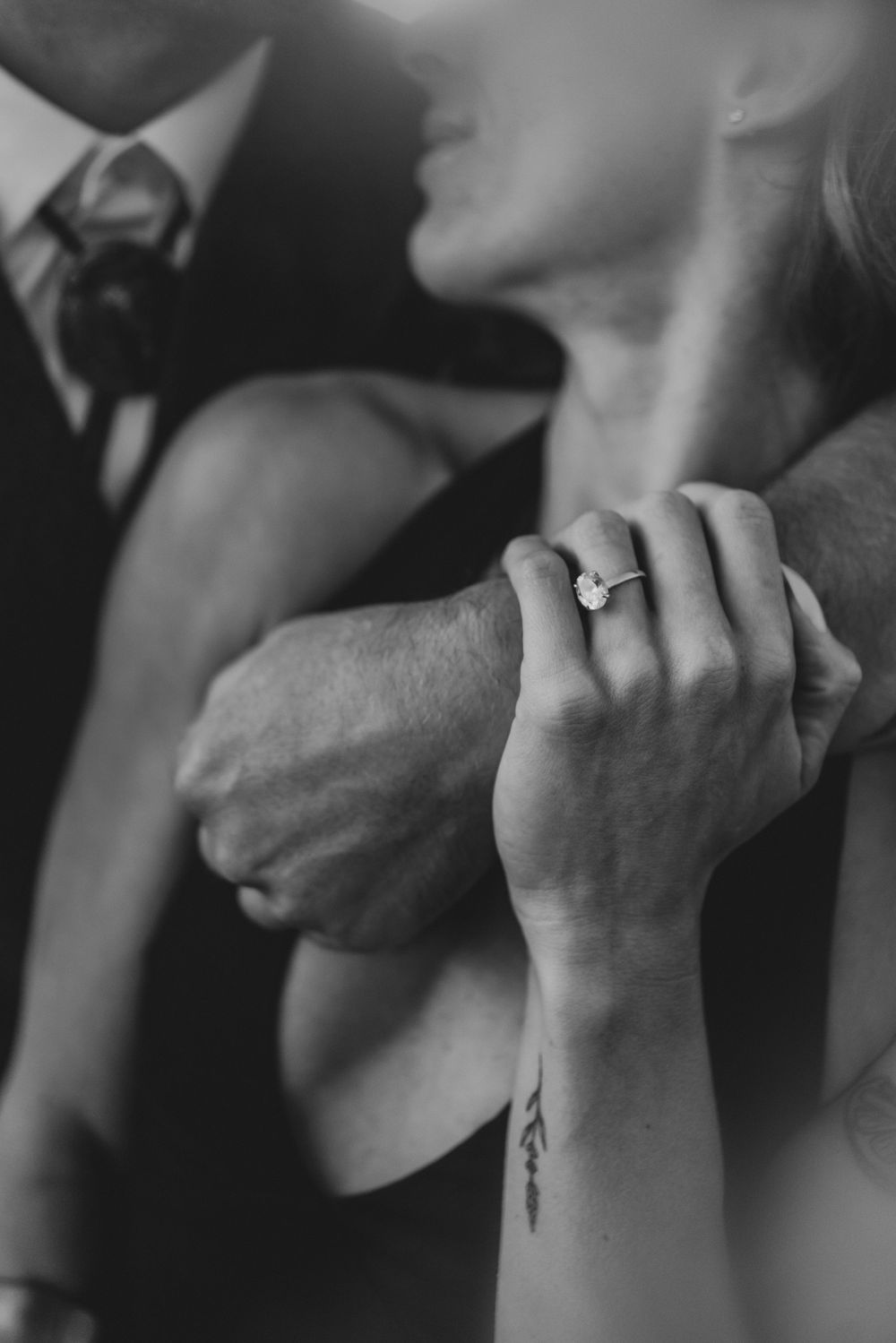 Black and white photo of a couple embracing, showcasing an elegant diamond engagement ring and a delicate wrist tattoo.