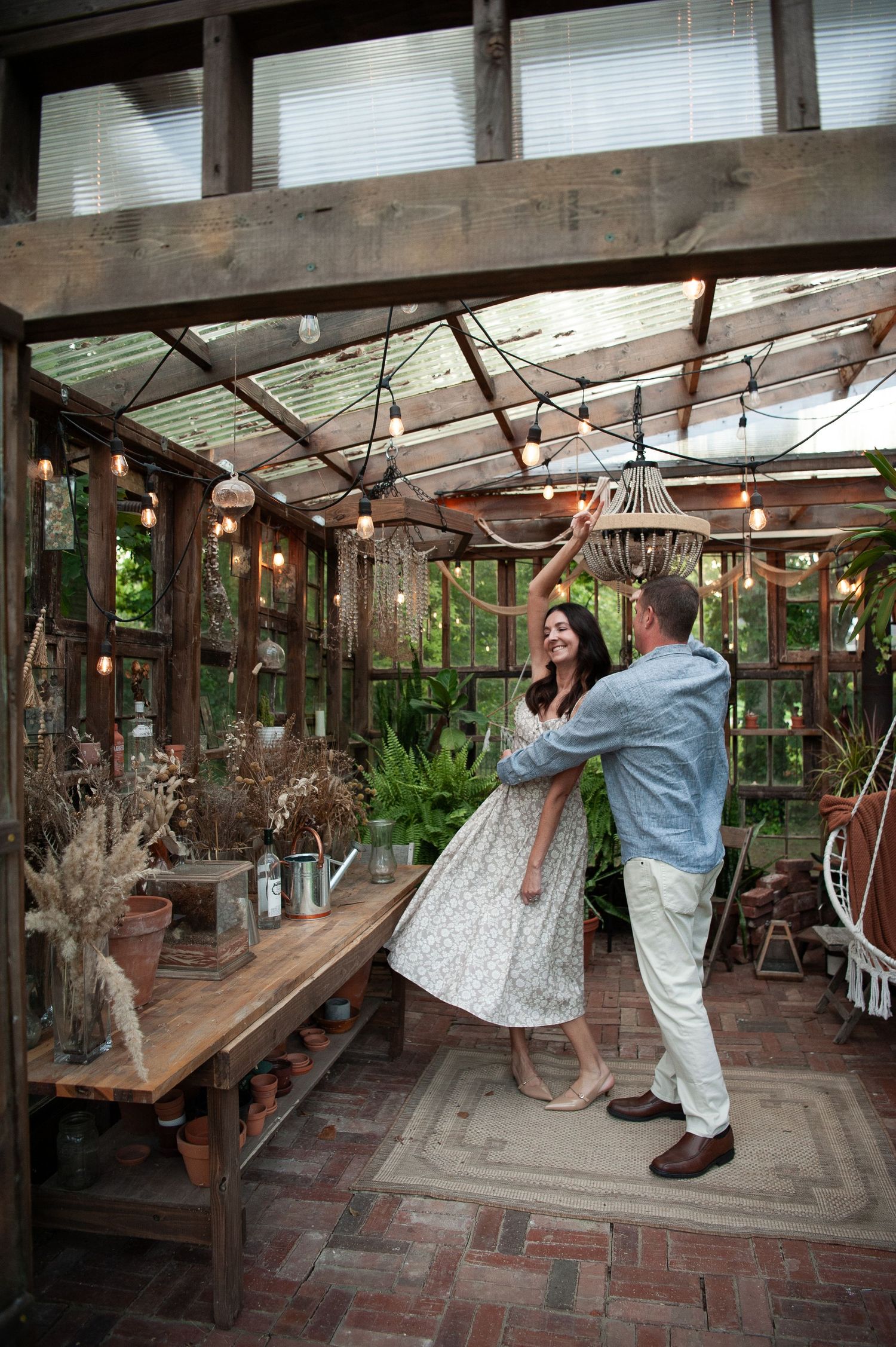 Couple dancing joyfully in a rustic greenhouse with string lights, brick floors, and lush plants surrounding them.