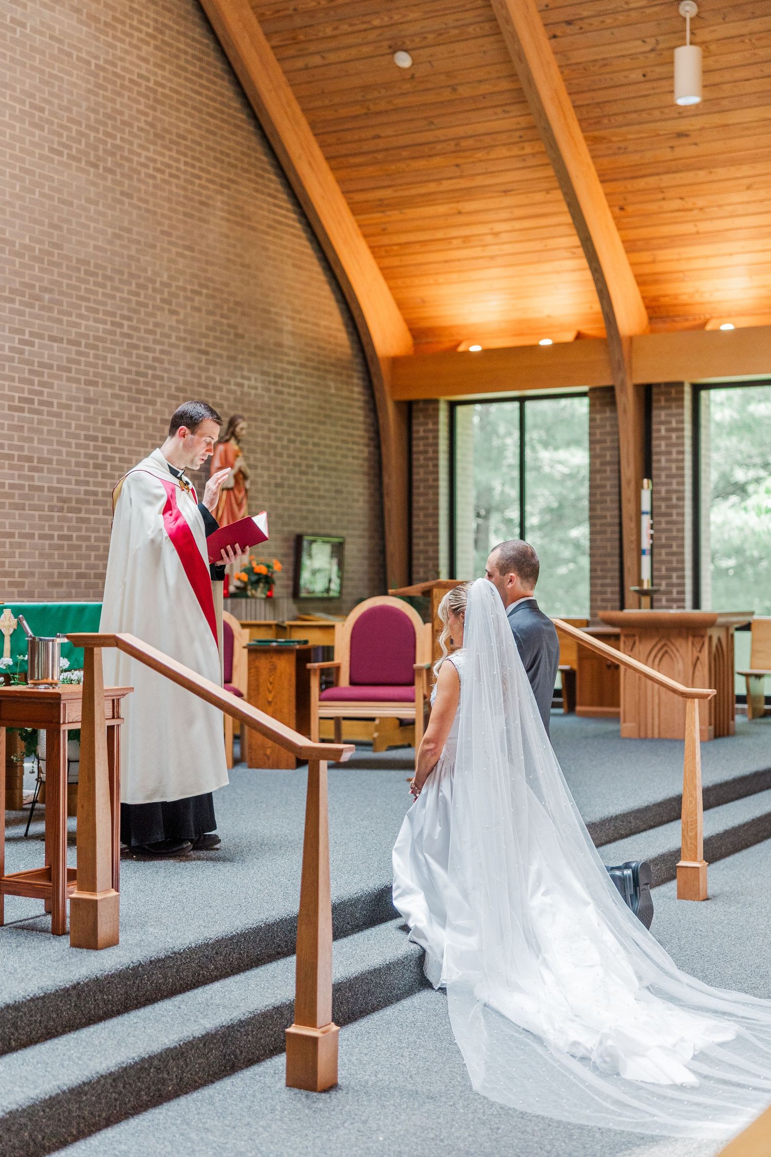 Bride and groom kneel before officiant during church wedding ceremony with wooden cathedral ceiling.