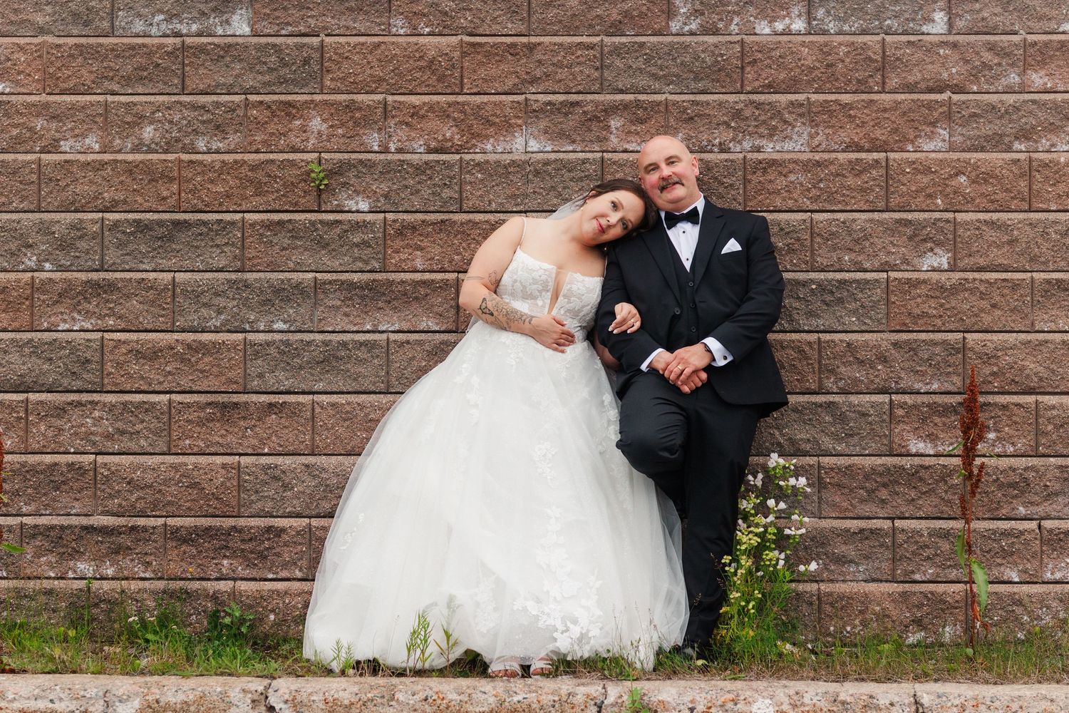 Bride and groom leaning against a brick wall, smiling in wedding attire, white ball gown and black tuxedo.