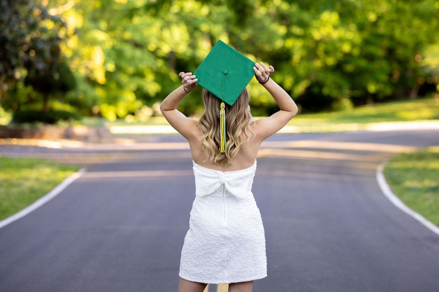 Smiling young woman in white dress holding navy graduation gown in sunlit meadow at golden hour.