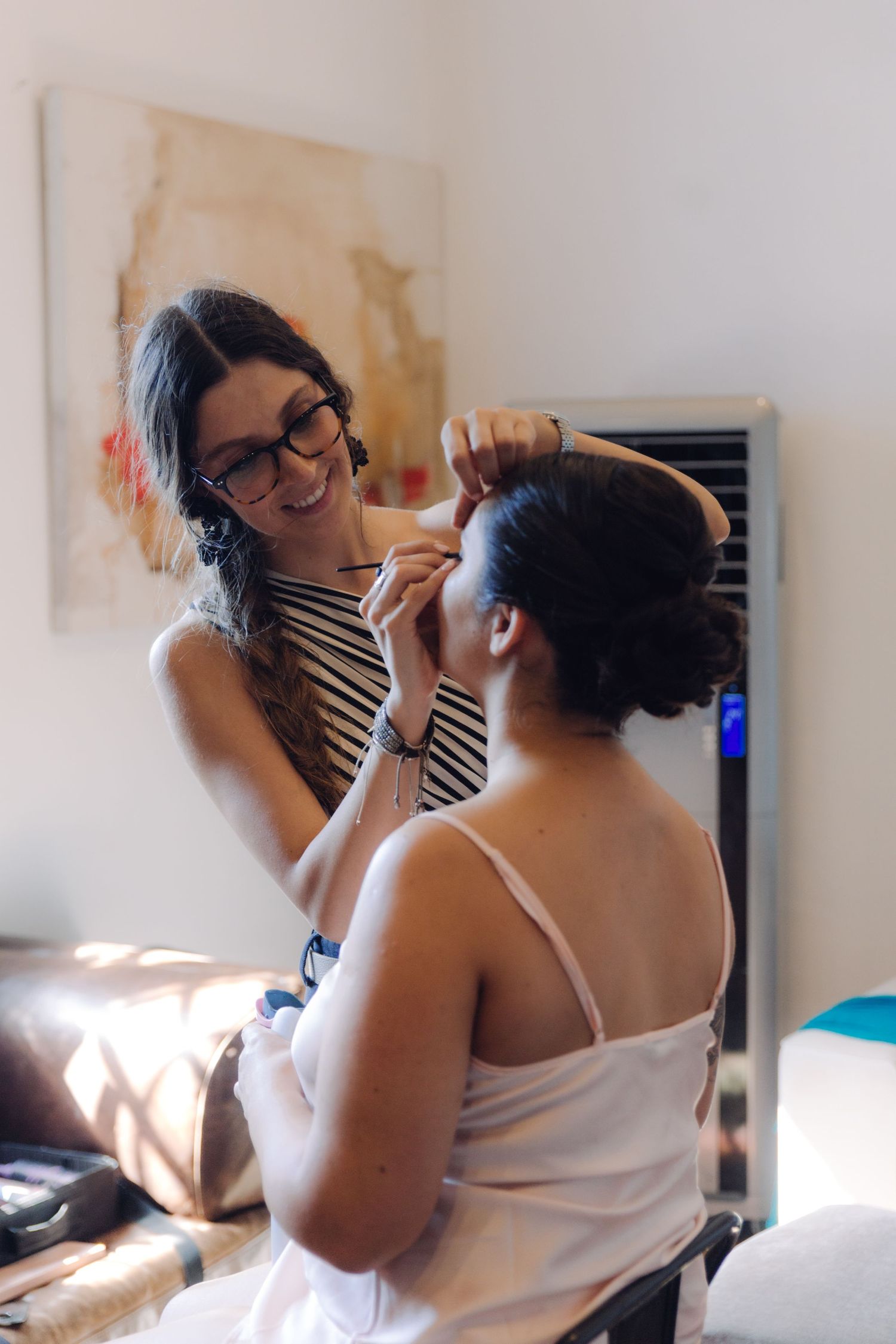 Smiling makeup artist styling bride's hair in bright room before wedding.