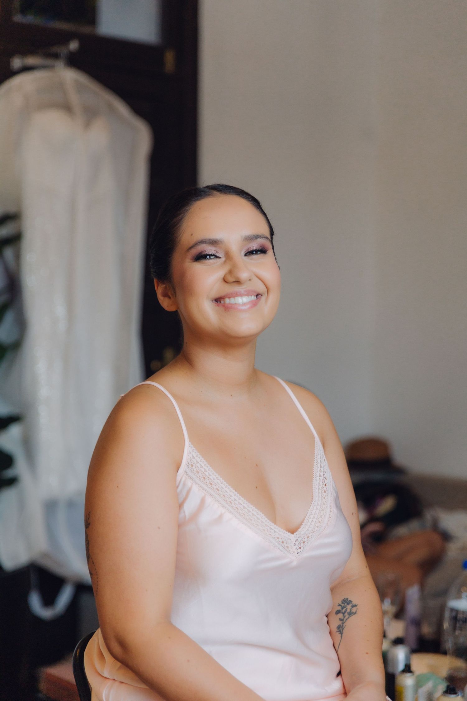Radiant bride in white slip dress smiling with wedding gown hanging behind her.