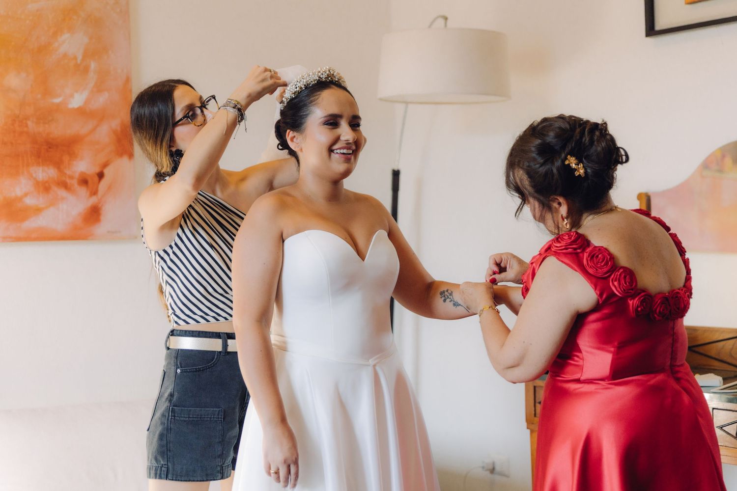 Bride in white gown being helped with tiara and dress by two women.