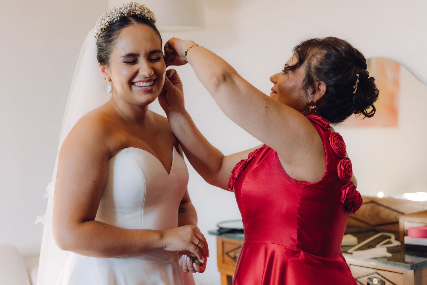 Mother in red dress helping bride with earrings during wedding preparations.