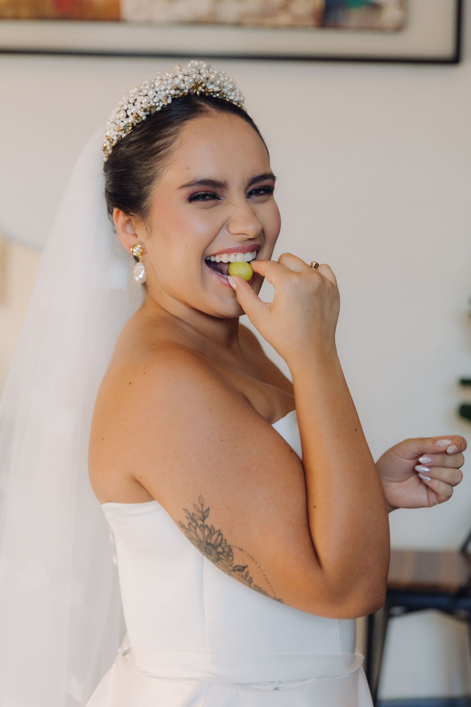 Joyful bride in white strapless gown and tiara eating a grape before ceremony.